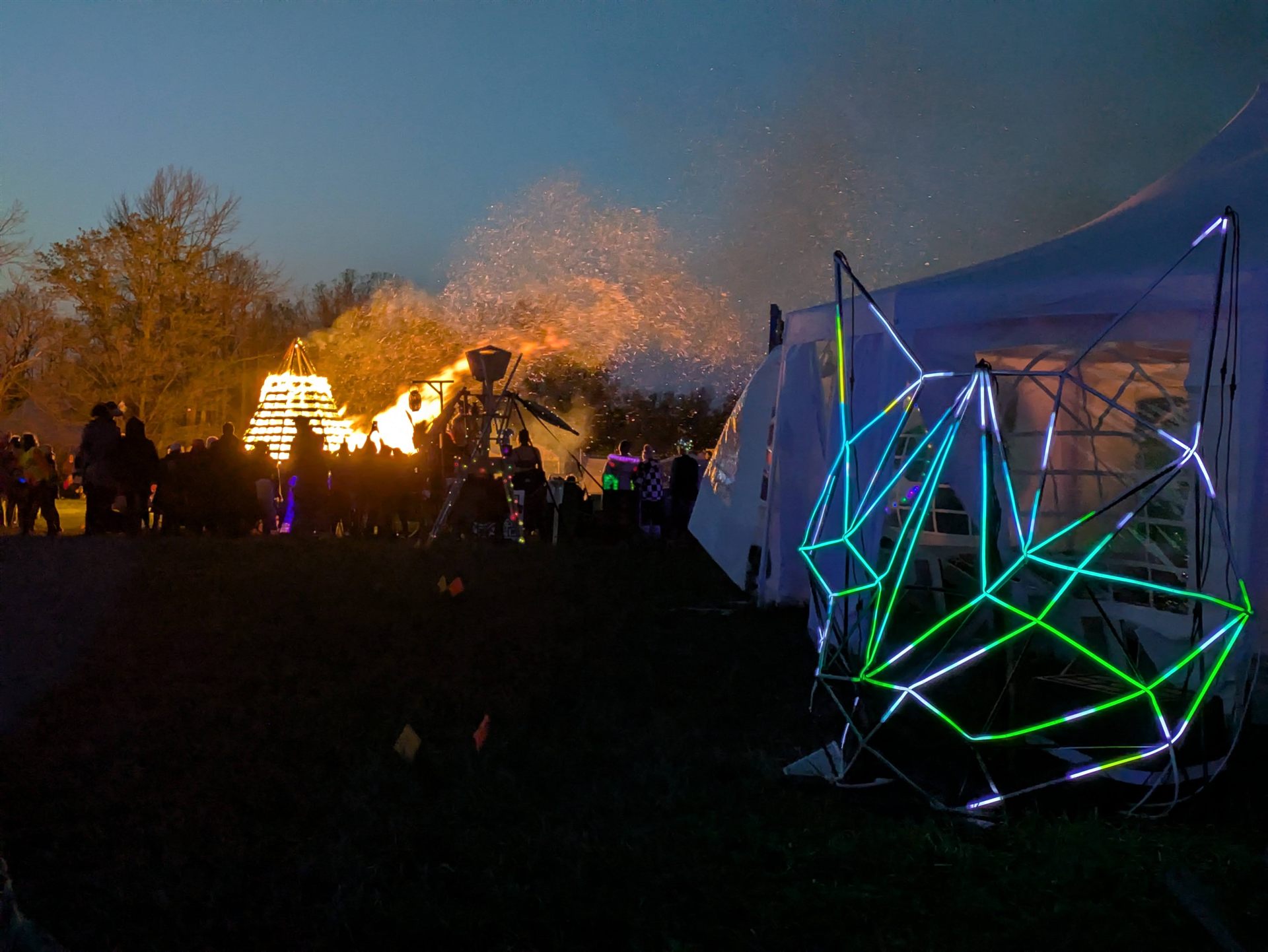 The sculpture lit green and blue beside a marquee at dusk, the SideBurn effigy alight in the distance.