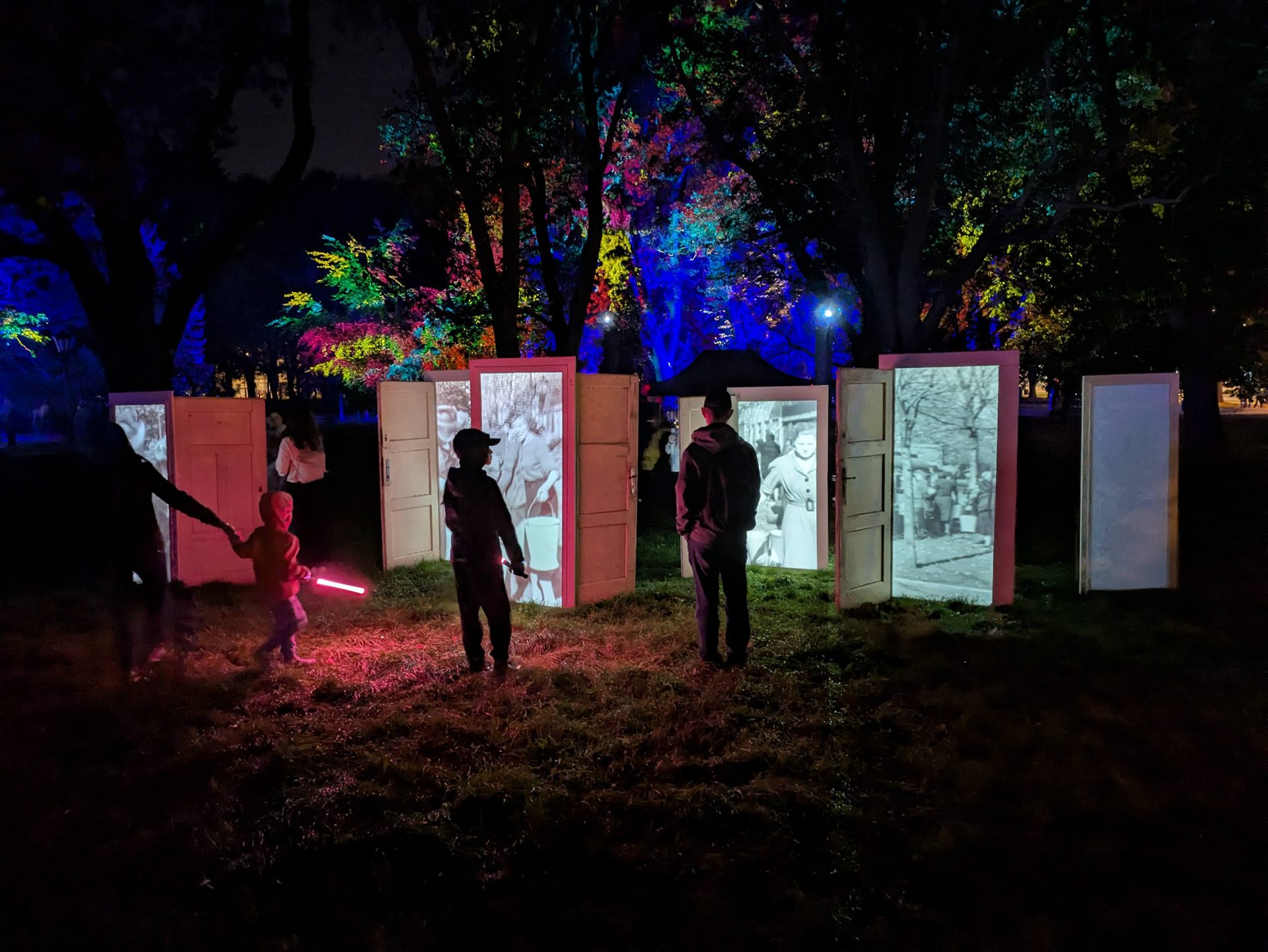 Visitors walking among the lit doors, park trees floodlit in blue and pink behind them.