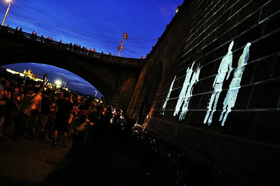 Figures projected onto the stone embankment wall at Náplavka at dusk, a crowd watching below and a bridge behind.